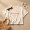 White t-shirt with 'sunkissed & unbothered' text, sunglasses, and seashells on a beige background