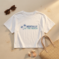 White t-shirt with 'Mentally at the Beach' text and palm tree design on sand with sunglasses and shells.
