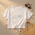 White t-shirt with 'Soft Life Beach Edition' text on sand with sunglasses, bag, and shells.