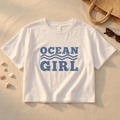 White t-shirt with 'Ocean Girl' text on sand with beach accessories