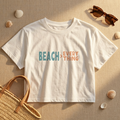 White t-shirt with 'BEACH > EVERYTHING' text on a sandy background with sunglasses, shells, and a bag.