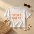 White t-shirt with 'Stay Salty' text, sunglasses, straw bag, and shells on a sandy background